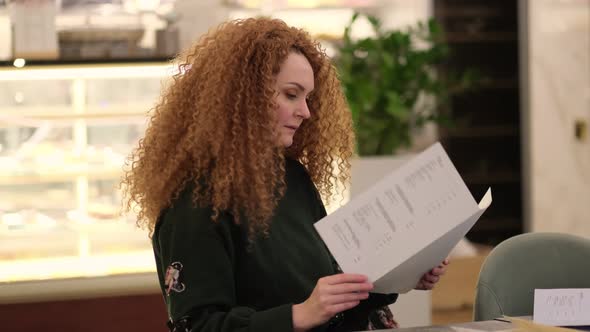 Bright Redhead Woman Sits at a Table in a Cafe and Chooses a Dish From the Menu