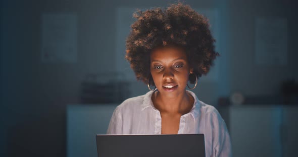 Portrait of Joyful Young Afroamerican Woman Sitting at Desk in Office Using Laptop Late in Evening