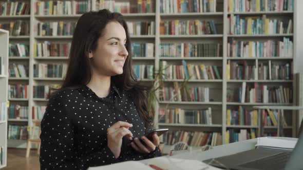 Confident Young Woman in Smart Casual Wear Holding Smart Phone and Looking at It with Smile While