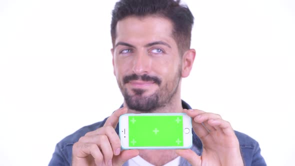 Face of Happy Young Bearded Hipster Man Thinking While Showing Phone