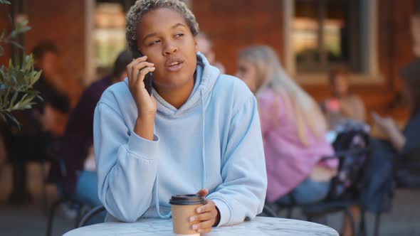 Young African Woman Talking on Smartphone Drinking Takeaway Coffee at Table Outdoors