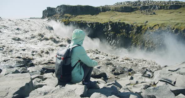 Female Hiker with Backpack Sitting at Cliff Edge Enjoying Detifoss Waterfall in Iceland