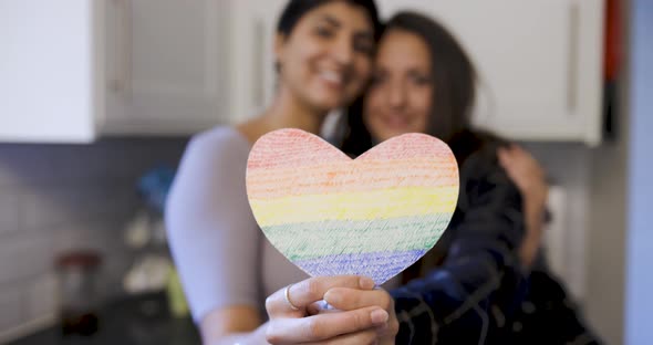 Happy girl friends lesbian couple embracing and holding a heart shaped rainbow flag