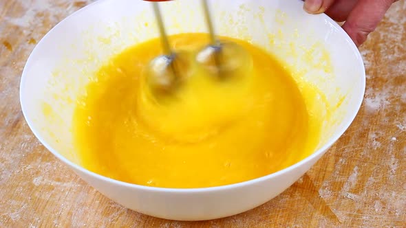 Woman Knocks Broken Eggs in a Large White Deep Dish with a Mixer on a Wooden Background Strewn with