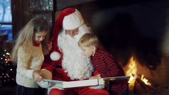 Children look into album in Santa's hands near fireplace.
