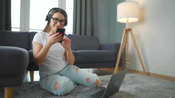 Casually Dressed Woman with Headphones is Sitting on Carpet with Laptop and Smartphone and Working