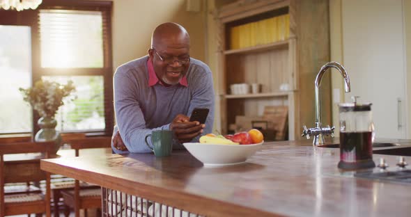 African american senior man leaning on kitchen counter using smartphone, taking off glasses, smiling