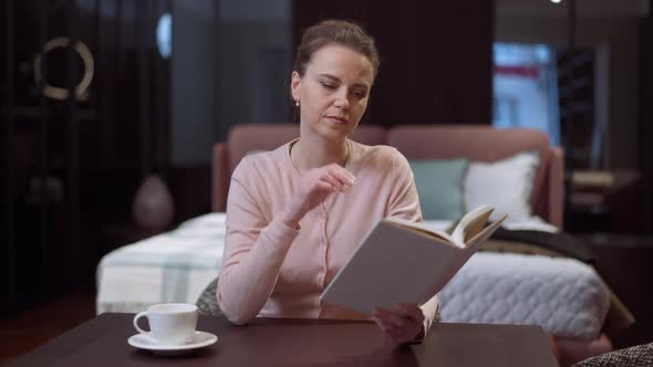 Portrait of Absorbed Caucasian Beautiful Slim Woman Sitting at Table in Bedroom Reading Book