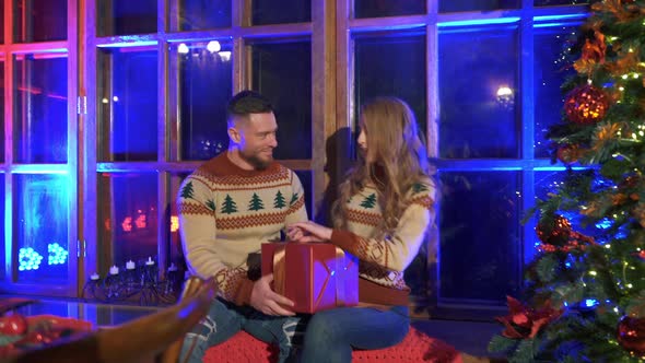 Newlyweds in knitted Christmas sweaters holding Christmas presents near Christmas tree.