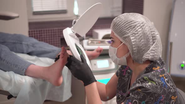 The Pedicure Master Looks at the Patient's Foot Through a Large Magnifying Glass