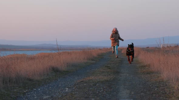 A Girl in a Jacket Runs and Fools Around on the Beach By the Sea with a Rottweiler Dog