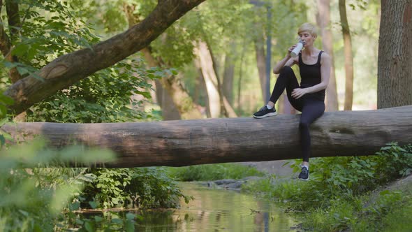 Slender Sportive Caucasian Sportswoman Woman Sitting on Log Tree in Park Outdoors in Green Forest