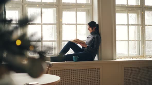 Smiling Young Woman Is Reading Book Sitting on Window-sill on Christmas Day and Enjoying Rest and