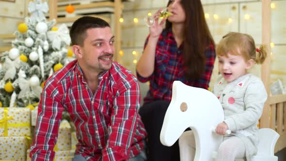 Little Girl Swings on Rocking Horse beside her Parents Celebrating Christmas and New Year at Home