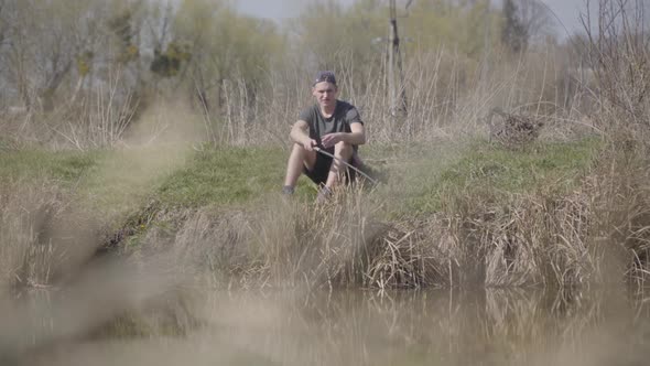 Wide Shot Portrait of Focused Young Caucasian Fisherman Sitting on Bushy Lake Shore and Looking at