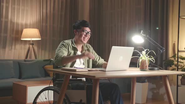 Asian Man Sitting In A Wheelchair While Video Call On Laptop Computer At Home, Night Time