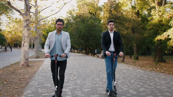 Two Handsome Young Businessman Riding an Electric Scooters in City Park During a Break and Talking