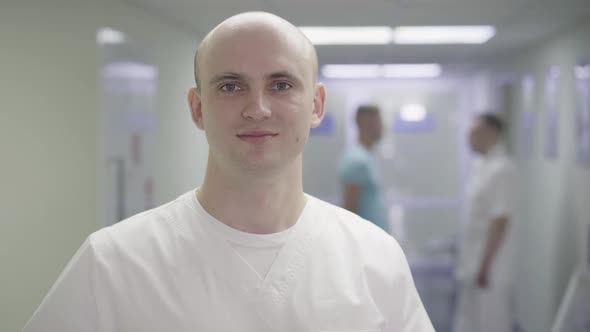 Close-up Face of Young Confident Caucasian Physician Posing in Hospital Corridor with Blurred People