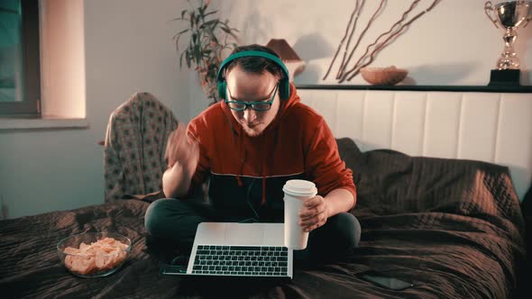 Young Man Sitting on the Bed and Talking with Somebody Using Laptop