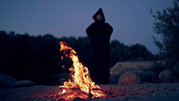 Powerful witch or wizard in black costume stands and watches the fire flame in the evening.