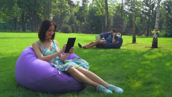 Young Brunette Girl Is Sitting in a Chair in the Park and Swipe Pages on Your Tablet