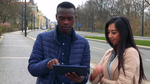 A Young Black Man and a Young Asian Woman Stand in a Street in an Urban Area and Work on the Tablet