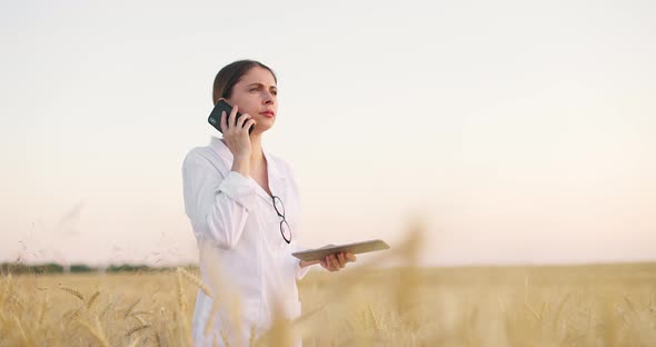 Young Caucasian Female Specialist Walking in Wheat Fieldchecking Harvest and Talking on Phone