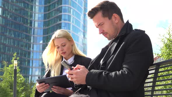 A Businessman and a Businesswoman Work on a Smartphone and a Tablet and Talk in an Urban Area