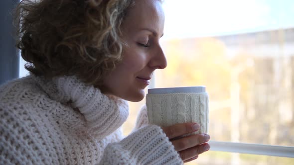 Young Woman In Knitted Sweater Smelling Tea Or Coffee Aroma By The Window