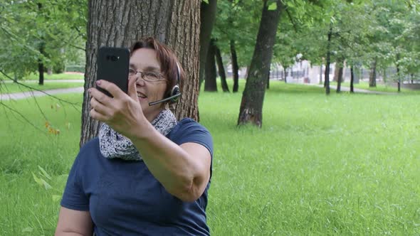 Senior Woman Having Video Call While Sitting on the Grass Under the Tree in City Park.