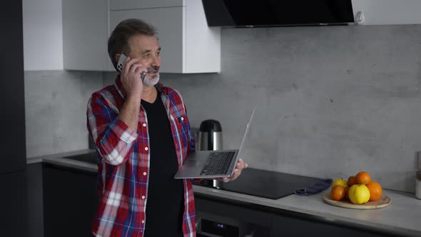 Modern Aged Man Using Laptop in Kitchen and Talking By Phone at the Same Time