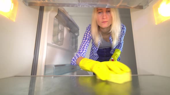 An inside view of a woman in rubber gloves cleaning an empty refrigerator.