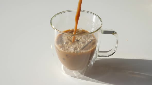 Fresh Coffee Pouring to Transparent Glass Cup with Ice on White Table Drops