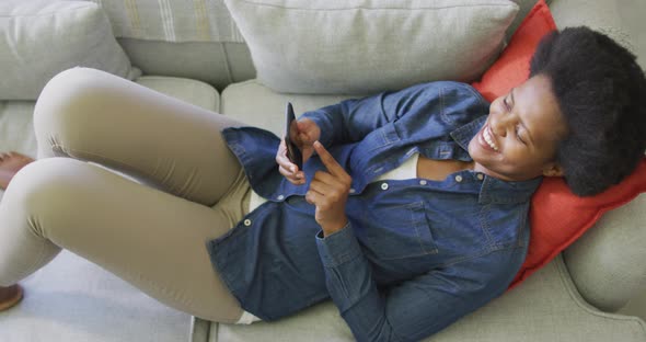 Happy african american woman laying on sofa, using smartphone