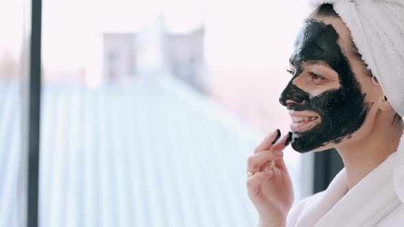 Pretty Woman with Wrapped Hair in a Towel Is Applying Black Mask on Face