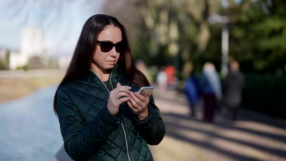 Brunette Woman with Sunglasses is Reading Messages in Her Smartphone Answering in Chat on Street