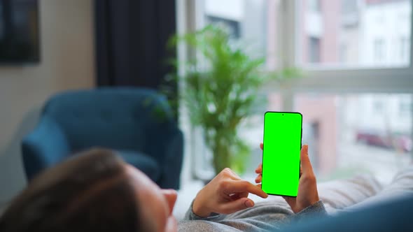Woman Lies at Home on the Sofa and Using Smartphone with Green Mockup Screen in Vertical Mode