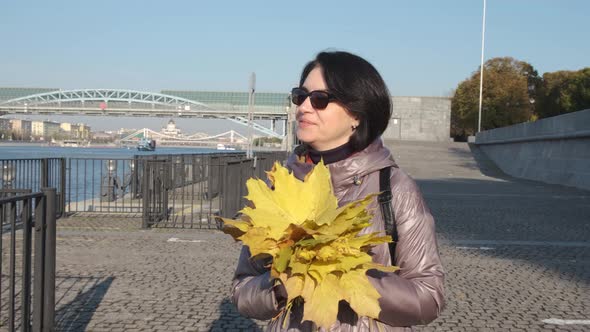 A Middleaged Woman Brunette with a Bouquet of Yellow Leaves Walks Along the City Embankment