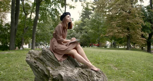 Girl Artist Sitting on a Large Stone in the Park with a Notebook in Hand