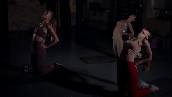 Three Flexible Fit Women Bending Back Standing on Knees on Exercise Mats in Darkness in Gym