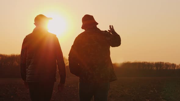 An Elderly and Young Farmer Go Together Over a Plowed Field at Sunset