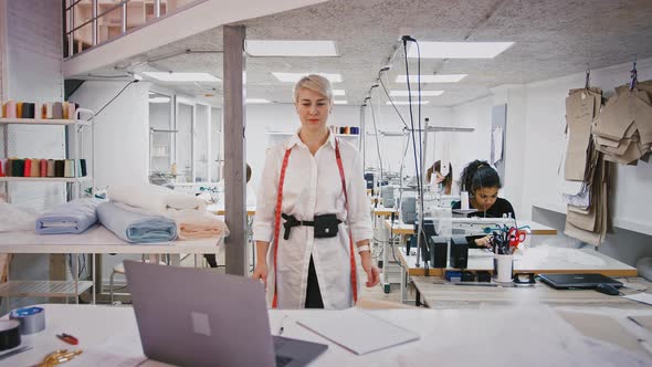 Woman Designer of Sewing Workshop Walking Over to Her Desk Watching Seamstresses at Work Sits Down
