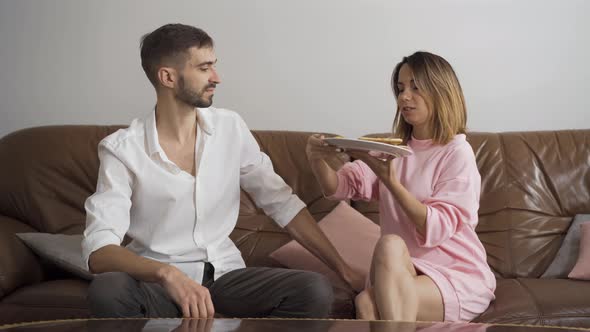 Woman in Casual Clothes Feeding Her Husband with Tasty Homemade Pizza Sitting on Leather Sofa. Happy