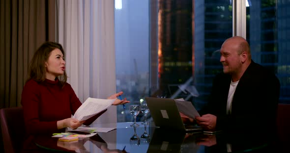 a Brunette and a Bald Middle-aged Man Sit at a Round Glass Table By a Large Window. They Hold