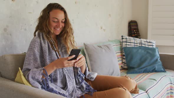 Smiling caucasian woman wearing blanket, sitting on sofa using smartphone in cottage living room