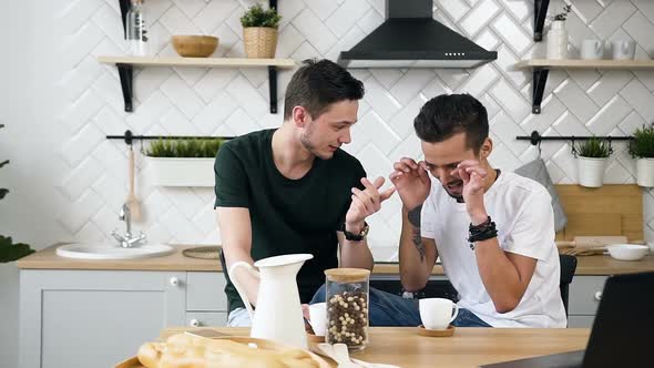 Gay Handsome Men are Sitting Behind the Kitchen Table Something Talking for During Drinking Coffee