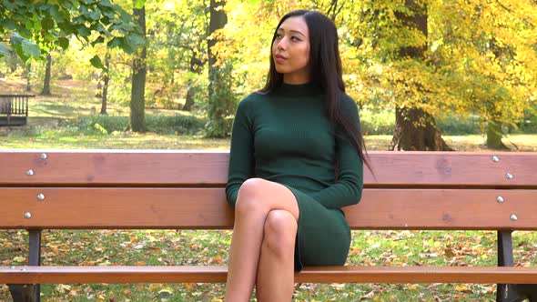 A Young Asian Woman Sits on A Bench in A Park and Looks Around