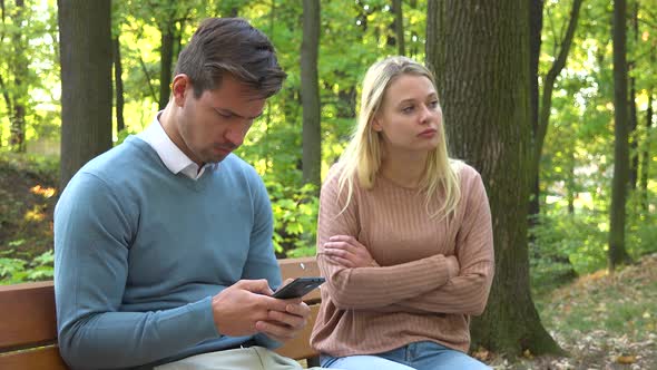 A Man and a Woman Sit in a Park, the Man Is Absorbed with His Smartphone, the Woman Is Angry at Him