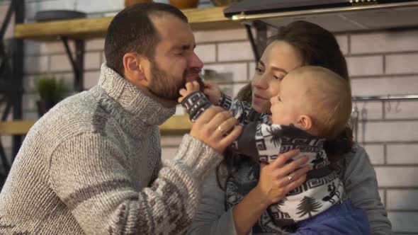 Smiling Father in Warm Sweater Playing with Baby Little Son Who Is in Mother Arms in the Kitchen