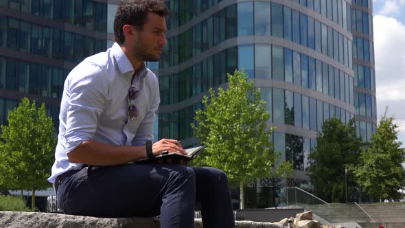 A Young Handsome Businessman Sits in a Park and Writes Notes - an Urban Area in the Background